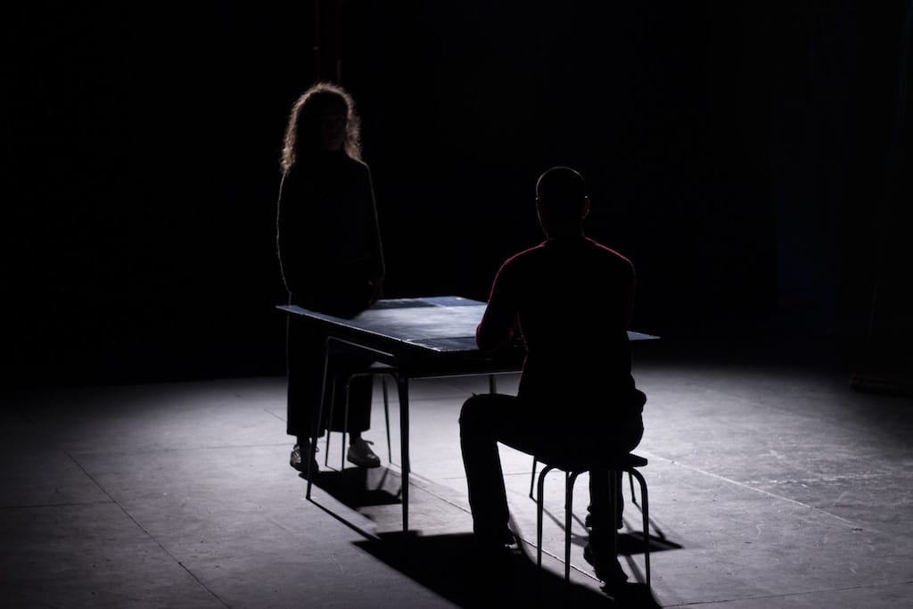 On a very dark stage, a man sits at a table and a woman stands across from him. The floor is illuminated but their faces are not visible.