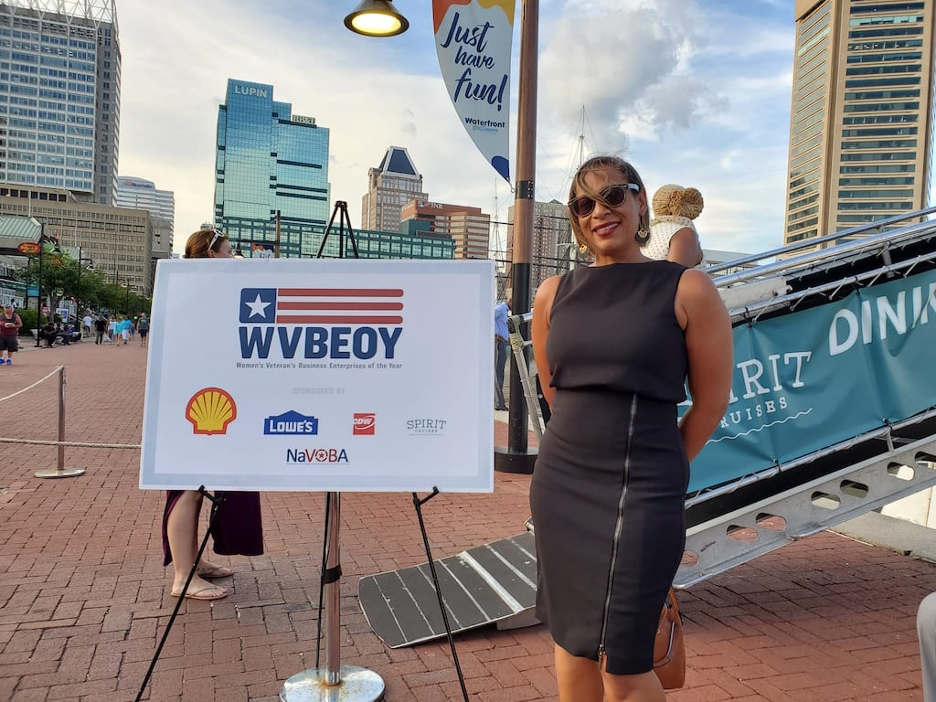 Woman in black dress and sunglasses standing to the right of a sign with the letters WVBEOY and logos in Baltimore's Inner Harbor.