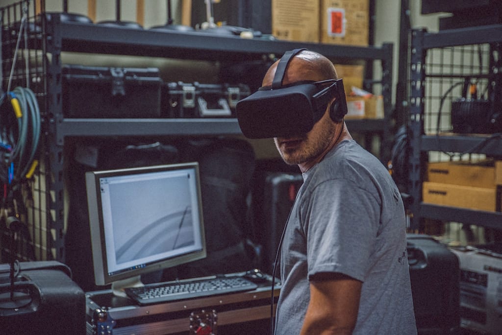 Man in t-shirt wearing virtual reality headset looking at the camera. Standing in front of a computer in an industrial warehouse space.