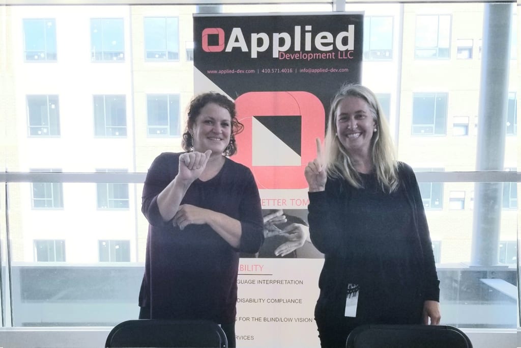 Two women standing inside in front of a sign that says "Applied Development." They are using American Sign Language to sign "Hi"