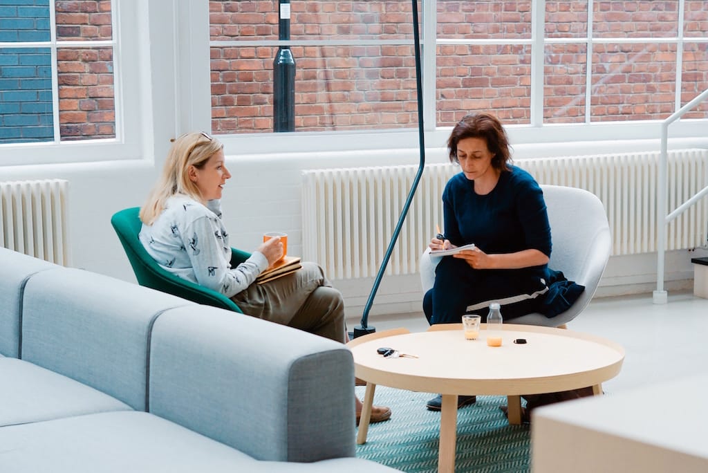 Two women sit opposite each other in an airy open office, one woman talks while the other takes notes.