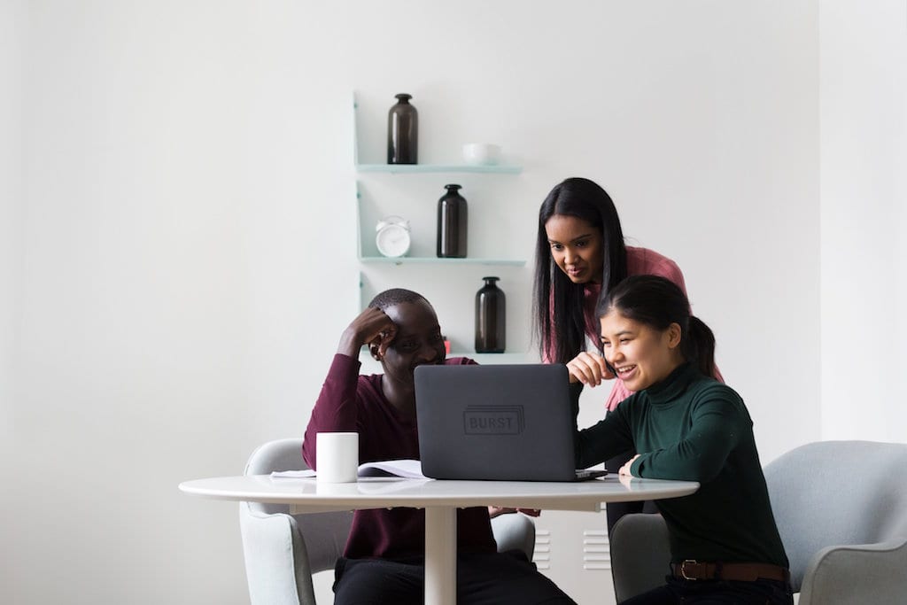 [Photo description: a man and a woman sitting at a table looking at a laptop computer screen with another woman standing looking at the screen. Photo by @sarahpflugphoto / via Burst Photo]