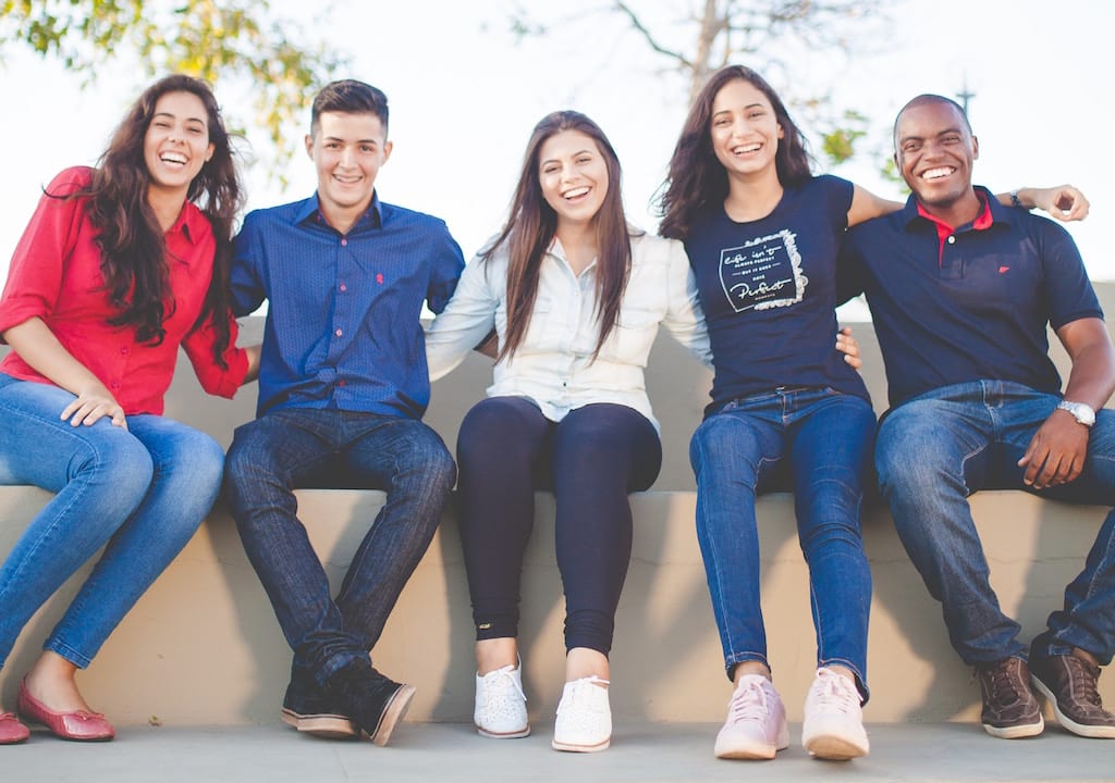 Five people sitting on a bench with their arms around each other smiling