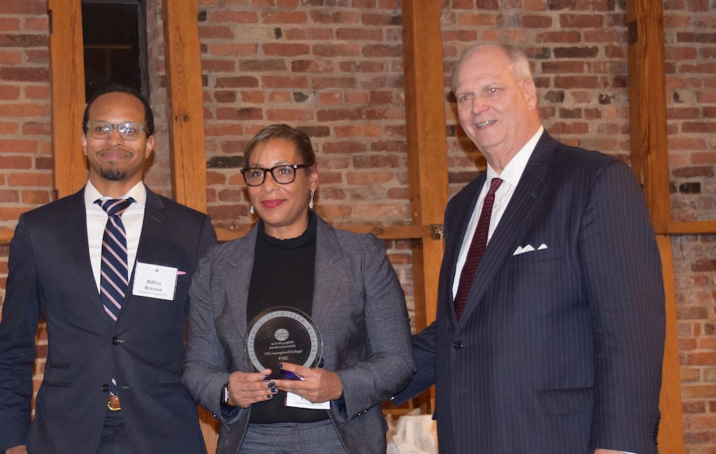 African American man with glasses, African American woman with glasses, and white man posing for the camera at a formal event. Woman holds an award.