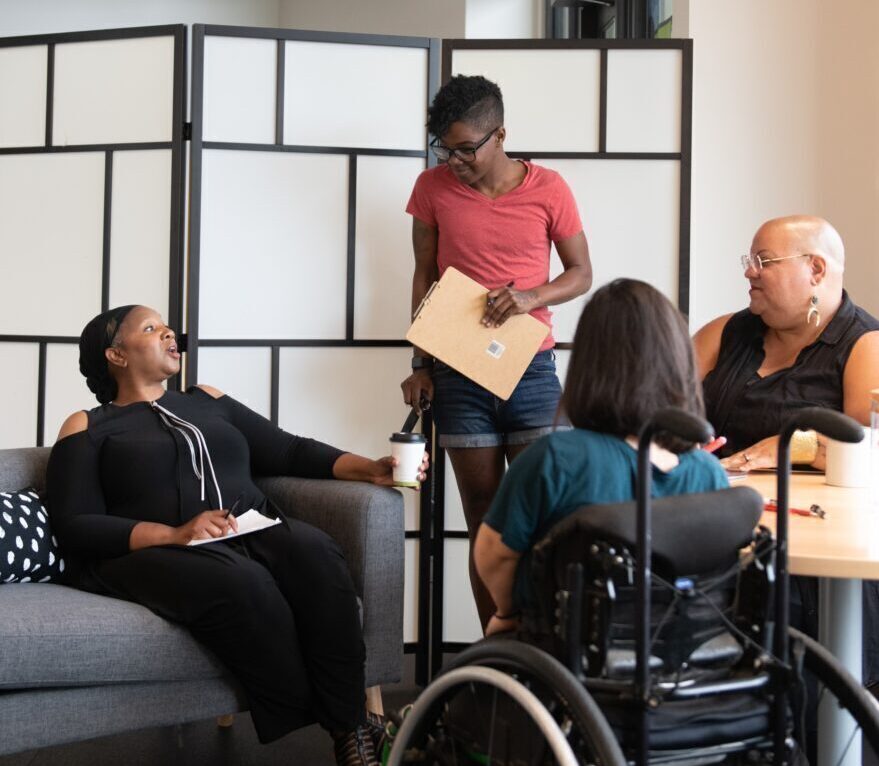 Four disabled people of color gather around a table during a meeting. A Black woman sitting on a couch gestures and speaks while the three others (a South Asian person sitting in a wheelchair, a Black non-binary person sitting in a chair, and a Black non-binary person standing with a clipboard and cane) face her and listen.