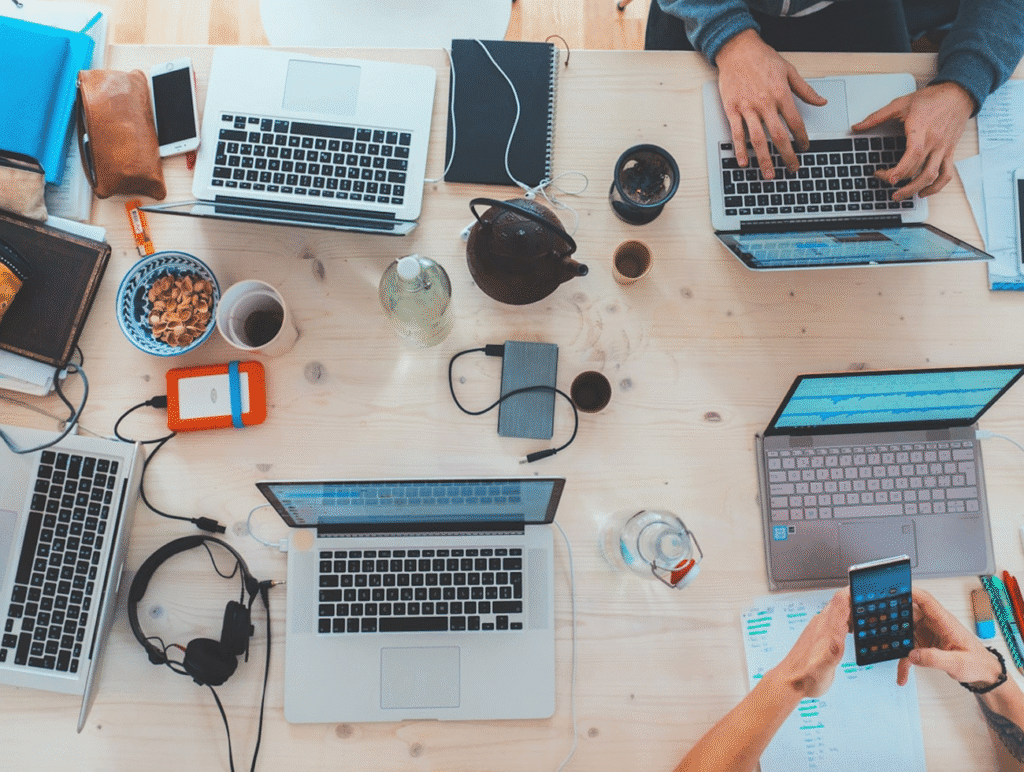 Overhead view of a busy workspace with laptops, smartphones, notebooks, and coffee, highlighting teamwork and technology.