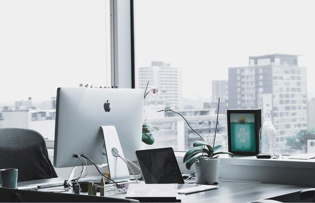 Modern office workspace with desktop computer, laptop, and a plant by a city view window.