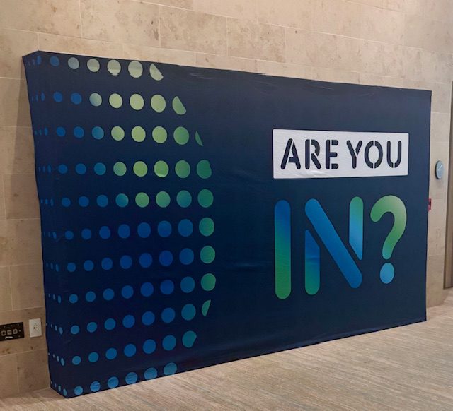 Banner with Are You In? text, featuring blue and green dots on a dark background, displayed in a modern space.