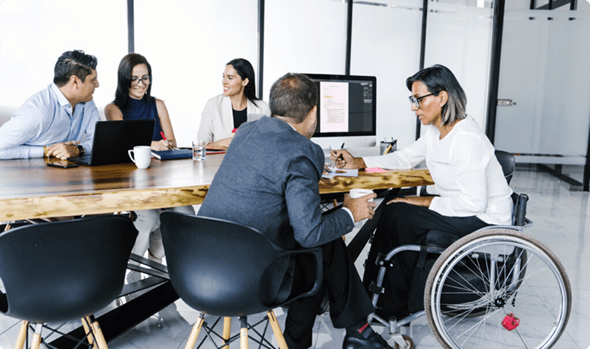 Diverse team in a modern office, engaged in a meeting around a wooden table, including a professional in a wheelchair.