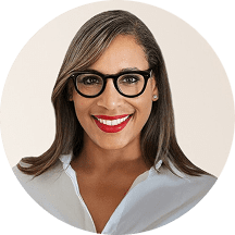 Smiling woman with glasses and red lipstick, wearing a white blouse, in front of a neutral background.