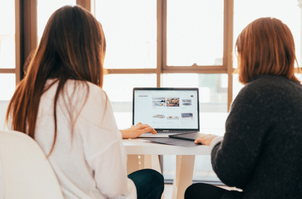 Two women working on a laptop in a bright room, viewing a website, collaborating at a modern workspace.