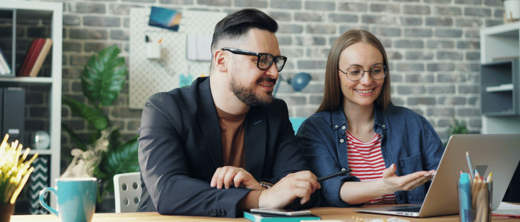 Two people collaborating in a modern office, looking at a laptop and discussing work with a brick wall backdrop.