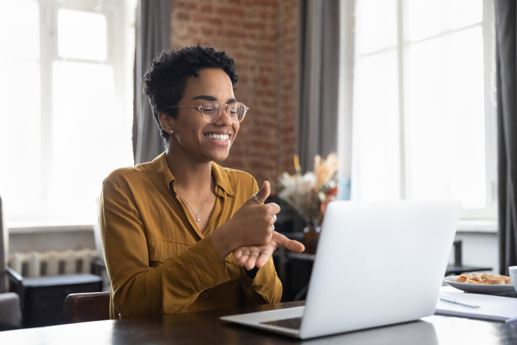 Smiling friendly African American therapist in glasses talking on video call, using sign language, speaking to patient with deafness, showing gestures at screen