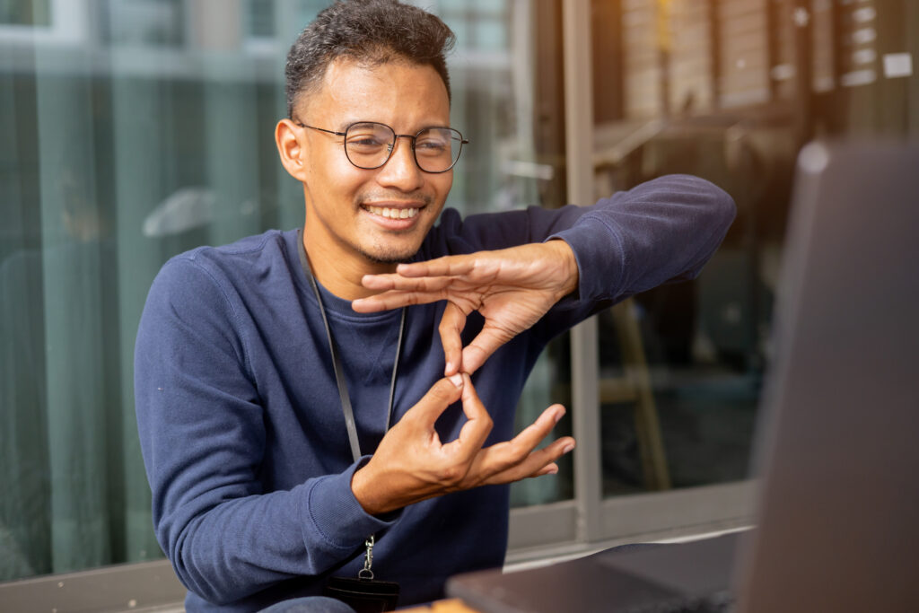 Man in glasses smiling while using sign language in a video call, sitting in front of a laptop.