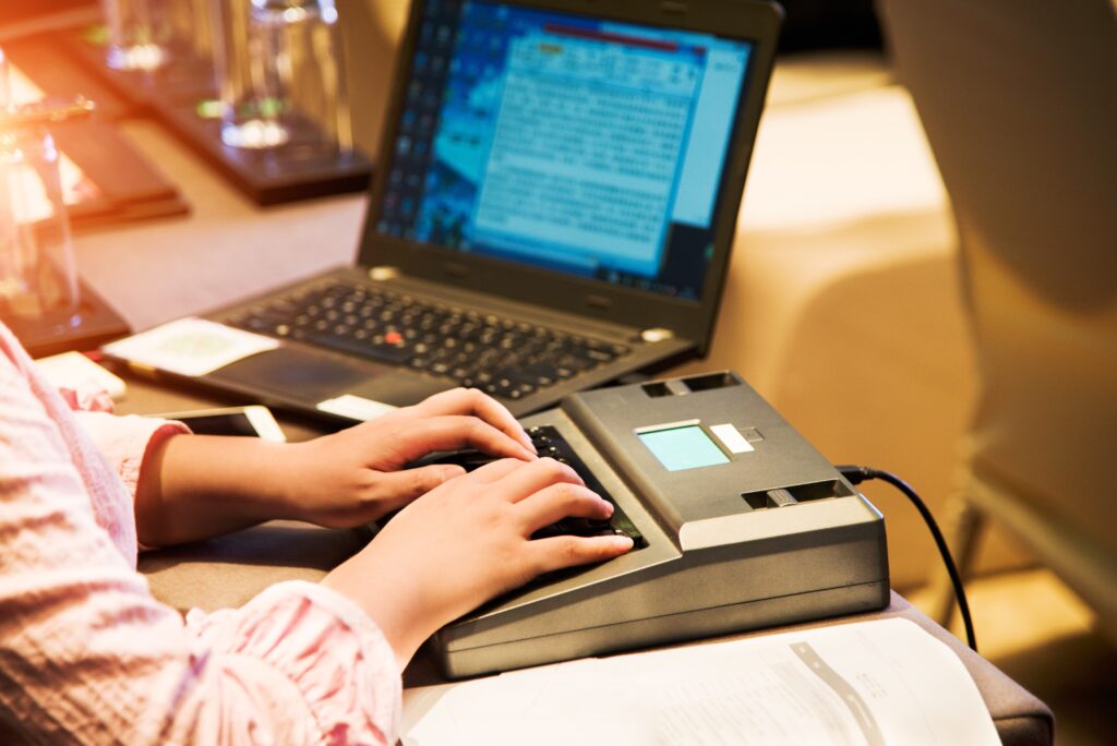 Person using a stenotype machine with a laptop, indicating transcription work or real-time captioning.