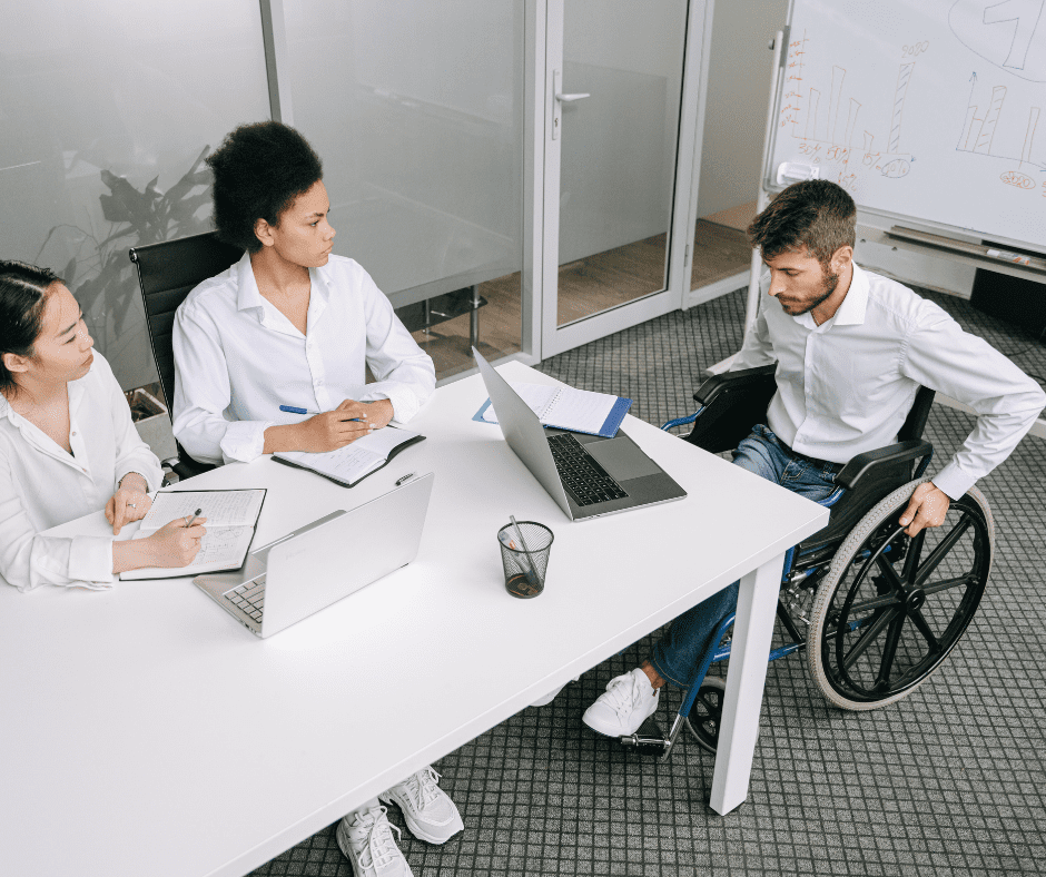 Team meeting in office with laptops and notes, including a person using a wheelchair. Inclusive workplace discussion.