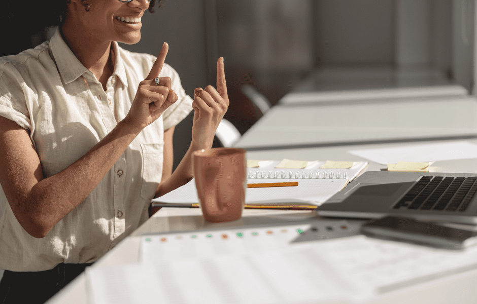 Person gesturing near a laptop and notebook in a bright office setting, conveying communication or learning.